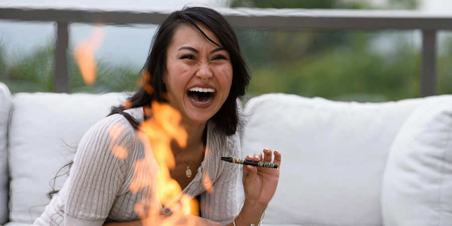 woman with black hair wearing white blouse laughing hard while holding The Clear CBD vape pen beside a bonfire outdoors